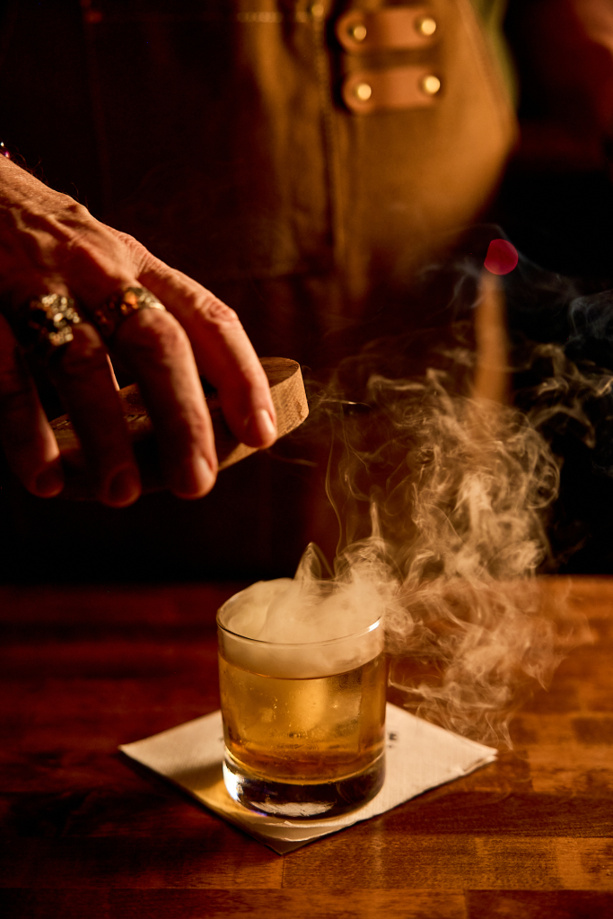 A bartender lifts a wooden smoke cover from a whiskey cocktail as smoke curls up from the glass