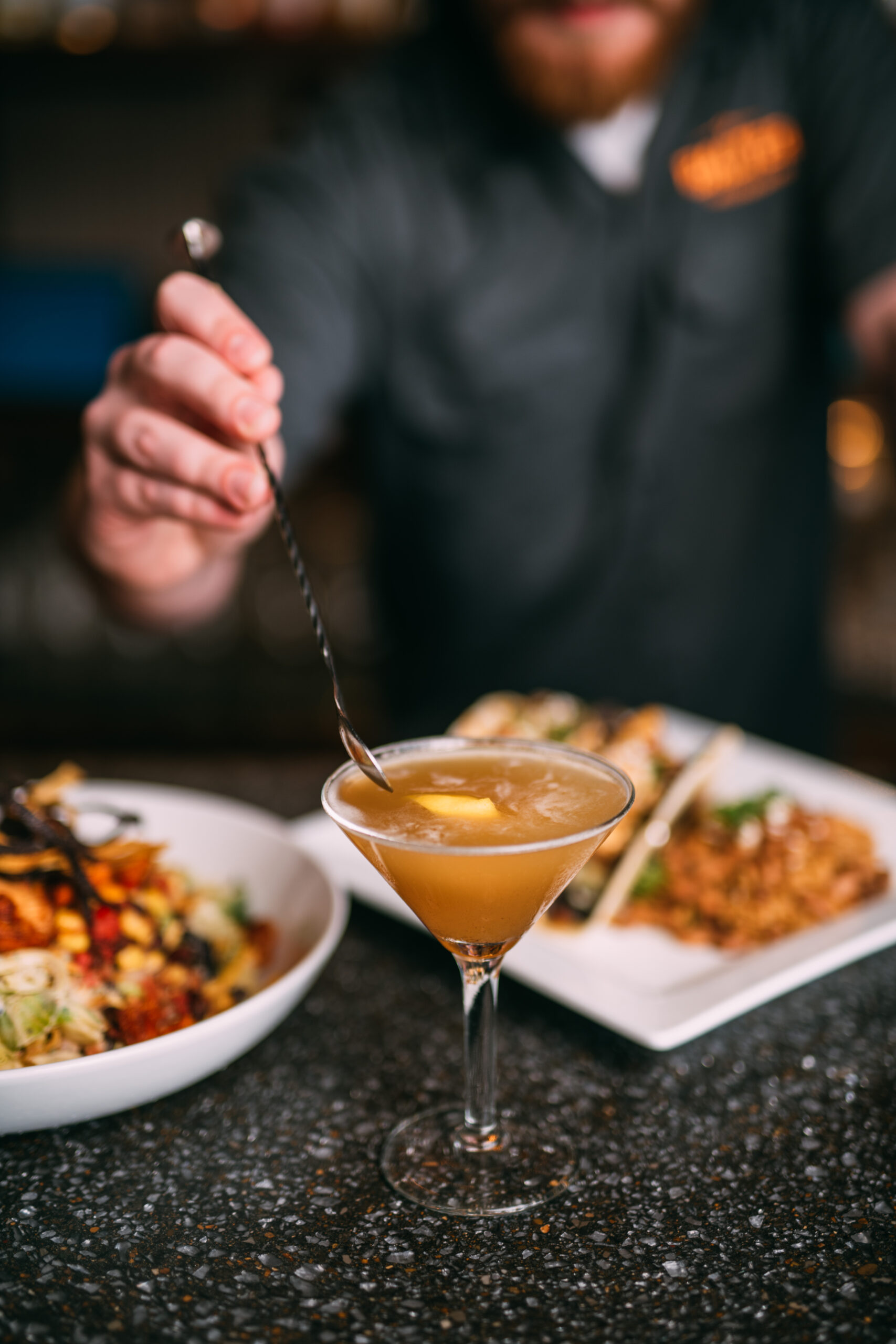 Bartender stirring a cocktail at a Tennessee Whiskey Week event with food in the background.